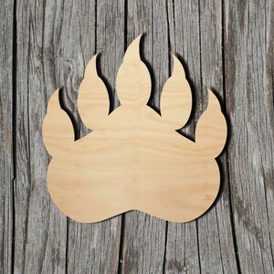 May include: A flat, light-coloured wooden cutout shaped like a bear paw print. The object features a smooth, natural wood grain surface with a distinct outline of four pointed claws and a large, rounded palm pad at the base of the design.