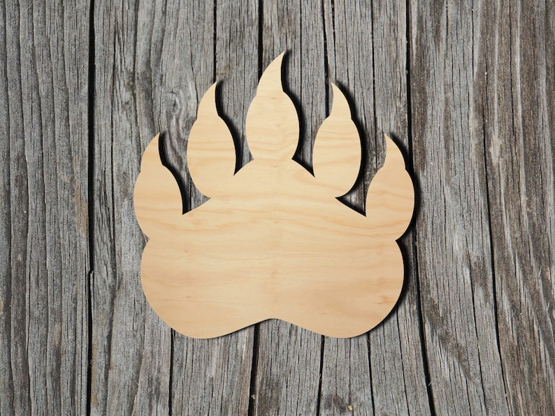 May include: A flat, light-coloured wooden cutout shaped like a bear paw print. The object features a smooth, natural wood grain surface with a distinct outline of four pointed claws and a large, rounded palm pad at the base of the design.