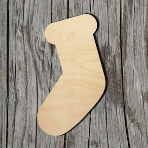 May include: A flat, unfinished light-colored wooden cutout in the shape of a Christmas stocking. The surface shows a natural wood grain pattern with a smooth, matte texture. The cutout features a curved toe and a folded cuff design.