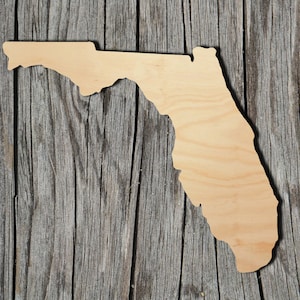 May include: A wooden cutout of the state of Florida, painted in a light wood color. The cutout is placed on a weathered wooden background.
