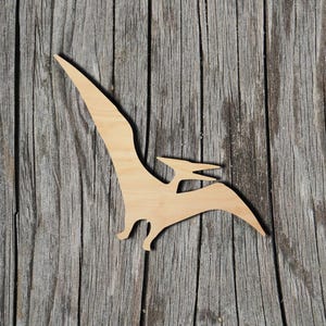 May include: A flat, light-colored wooden cutout in the shape of a pterodactyl dinosaur. The silhouette features a long, pointed wing extending upward, a smaller head with a beak, and a tail section, showcasing the natural grain of the wood.