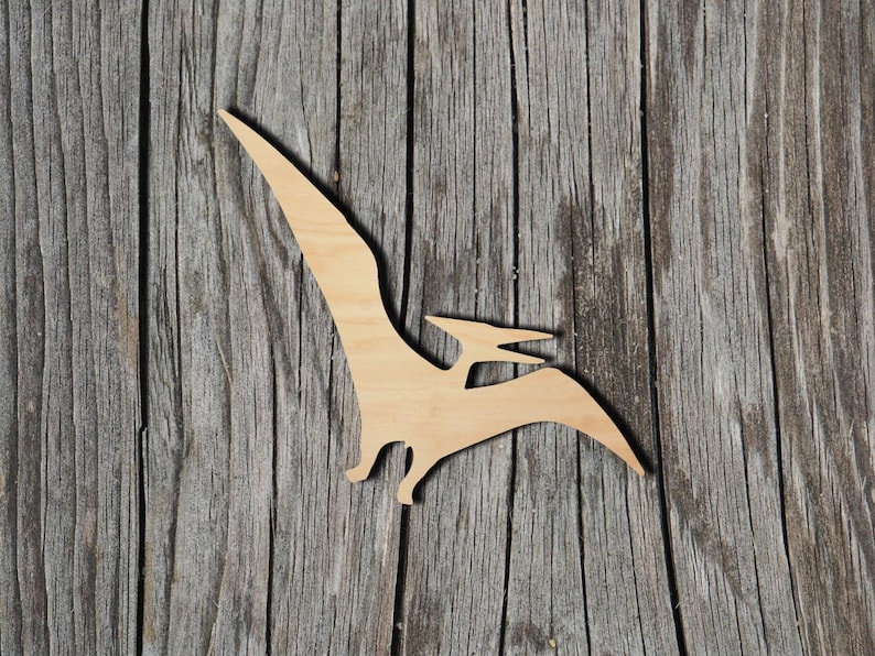 May include: A flat, light-colored wooden cutout in the shape of a pterodactyl dinosaur. The silhouette features a long, pointed wing extending upward, a smaller head with a beak, and a tail section, showcasing the natural grain of the wood.