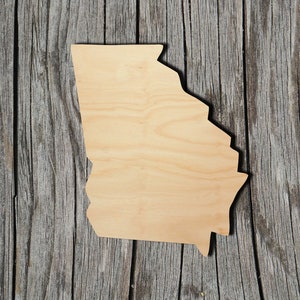 May include: A wooden cutout of the state of Georgia. The cutout is light brown and has a smooth surface. It is placed on a weathered wooden background.