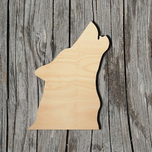 May include: A flat, light-colored wooden cutout shaped like the silhouette of a howling wolf. The wood features a natural grain pattern with visible light brown tones, showcasing a smooth surface and a distinct profile of a wolf head and neck.