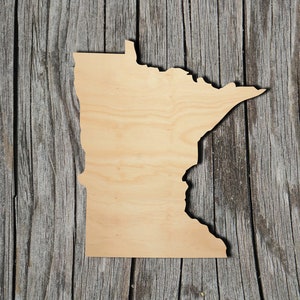 May include: A wooden cutout of the state of Minnesota. The cutout is light brown and has a smooth surface. It is placed on a weathered wooden background.