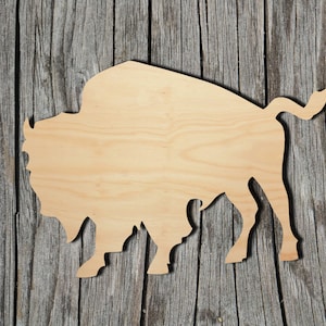 May include: A wooden cutout of a bison silhouette. The cutout is made of light brown wood and is set against a rustic wooden background.