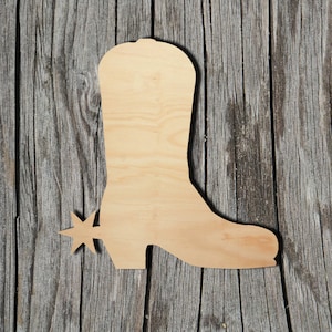 May include: A wooden cutout of a cowboy boot with a spur. The boot is light brown and has a detailed shape, including the heel, shaft, and toe. The spur is attached to the heel. The cutout is placed on a weathered wooden surface.