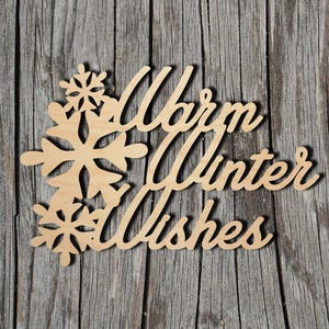 May include: A wooden cutout with the words "Warm Winter Wishes" in a cursive font, accompanied by snowflake designs. The cutout is made of light-colored wood and rests on a weathered, gray wooden surface.