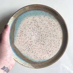 May include: A handmade ceramic plate with a speckled off-white center, a light blue ring, and a dark brown rim. The plate is round and has a rustic, artisanal appearance, perfect for serving food.