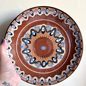 Peut inclure: Une assiette décorative en céramique avec une base marron et des motifs complexes en blanc, noir et bleu. L'assiette présente un motif central avec des cercles concentriques et un motif répétitif sur le bord. L'assiette est tenue par une main.
