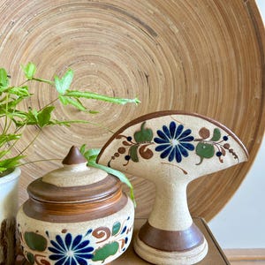 May include: A ceramic jar with a lid and a decorative fan-shaped piece. Both are beige with blue floral designs and green accents. The jar is round, and the fan-shaped piece has a pedestal base.