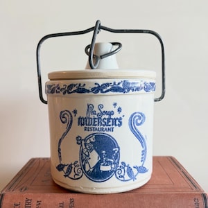 Peut inclure: Un pot en céramique crème avec un motif bleu imprimé représentant le texte Pea Soup Andersen's Restaurant, une illustration de chef et des volutes décoratives. Il comprend un couvercle assorti et une anse en fil de métal foncé.