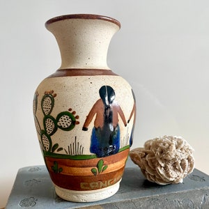 May include: A small, white ceramic vase with a brown rim and a painted design of two figures and a cactus. The vase has a brown, orange, and green striped design at the bottom and the word "CONC" is visible.