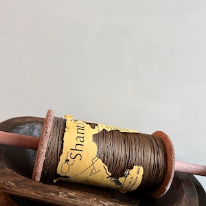 May include: A brown thread spool with a yellow label that reads "Shanti". The spool is resting on a wooden dowel and a wooden bowl. The thread is tightly wound around the spool.
