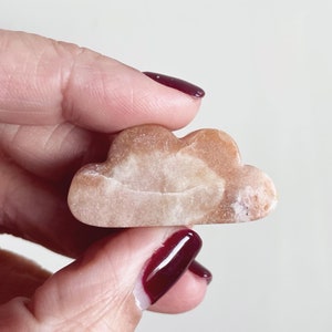 May include: A light brown cloud-shaped stone with white and brown marbling. The stone is being held by a hand with dark red nail polish.