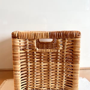 May include: A square, woven wicker basket with a natural, light brown color. The basket has a rectangular opening on the top and a handle cut out on the front. The basket is designed for storage and organization.