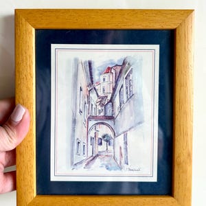 May include: Watercolor painting of a European street scene in a wooden frame. The artwork depicts a narrow alleyway with buildings on either side, an archway, and a small tree. The painting is set against a dark blue mat.