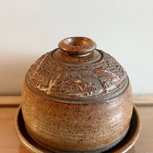 May include: A handcrafted, speckled brown ceramic butter dish with a lid. The lid features a detailed, textured design and a small, circular handle. The dish sits on a matching plate, creating a cohesive and rustic aesthetic.