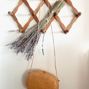 May include: A woven, oval-shaped, light brown handbag with a long, brown strap hanging from a wooden coat rack with dried lavender flowers.