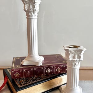 May include: Two white, classical pillar candle holders, one tall and one short. They are placed on top of two antique books with gold-gilded pages and decorative covers. The candle holders have a detailed, ornate design, reminiscent of ancient architecture.