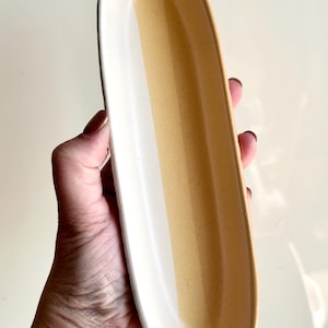 May include: A long, ceramic serving dish with a white and tan colour scheme. The dish is held in a hand and has a curved, oblong shape. The tan section is on the right side of the dish.