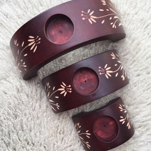May include: Set of three wooden candle holders in a graduated size. Each holder is a half-circle shape with a single candle well and a floral design carved into the wood. The holders are stained a dark reddish-brown color.