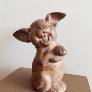 May include: A brown ceramic rabbit figurine with a wide, toothy grin. The rabbit is standing on its hind legs with its front paws raised. The figurine has a rustic, hand-painted look.