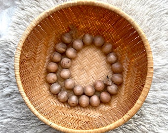 May include: A woven basket with a natural wood finish holds a circle of wooden beads. The beads are a light brown color and are arranged in a circle.
