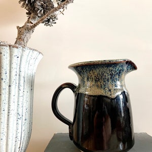 Peut inclure: Pichet en céramique fait à la main, avec une base marron foncé et un bord bleu et beige. Le pichet a une anse incurvée et un bec verseur. Un vase blanc et bleu et une branche avec des pommes de pin sont en arrière-plan.