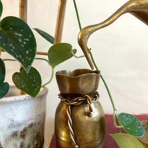 Peut inclure: Petit vase en laiton antique avec un design unique de corde nouée au milieu. Le vase a un dessus à volants et repose sur un livre rouge. Des plantes vertes et une statue de grue dorée sont en arrière-plan.