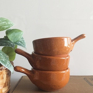 Può includere: Tre ciotole in ceramica marrone con manici impilate l'una sull'altra. Le ciotole sono smaltate e hanno un aspetto rustico.