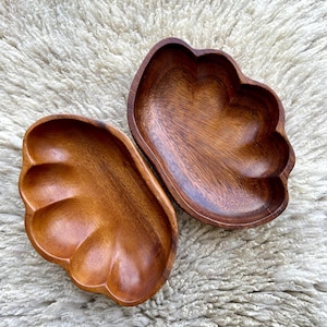 Può includere: Due ciotole in legno a forma di foglia, di un ricco colore marrone caldo. Le ciotole hanno una finitura liscia e lucida e sono poste su uno sfondo strutturato bianco sporco. Le ciotole sono vuote e sembrano fatte a mano.