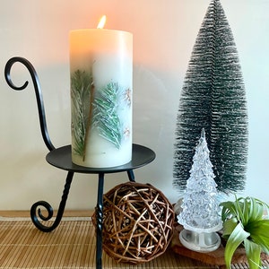 May include: A lit, white pillar candle with a pine needle and pine cone design sits in a black metal candle holder. A small, green bottle brush tree and a clear, plastic Christmas tree are also in the image, along with a woven ball.