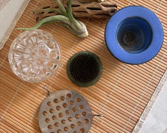 May include: A collection of floral arrangement tools including a clear glass flower frog with a textured surface, a round metal pin frog with dark needles, a circular silver-toned metal grid with wire supports, and a blue ceramic vessel.