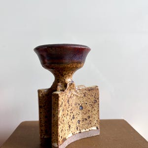 Peut inclure: Un vase en céramique au design unique. Il présente un dessus en forme de bol bordeaux foncé, un corps beige moucheté et une base incurvée. Le vase est un objet décoratif.