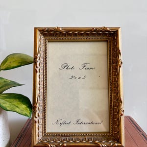 Puede incluir: Un marco de fotos rectangular de tono dorado con un borde texturizado y ornamentado. El papel insertado muestra el texto "Photo Frame 3 1/2 x 5" y "Norfleet International" en letra negra. El marco es para una fotografía de 8,9 cm por 12,7 cm.