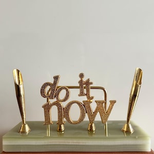 May include: A gold desk tidy with the words "do it now" in a decorative font. The organiser includes two pen holders and sits on a light green and white stone base.