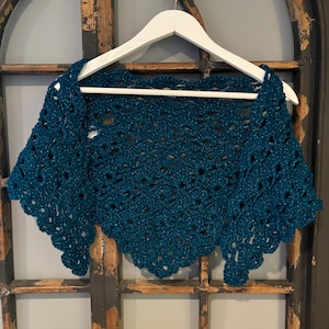 Puede incluir: Chale de ganchillo de color turquesa con un patrón de encaje, colgado de una percha blanca sobre un fondo de madera.