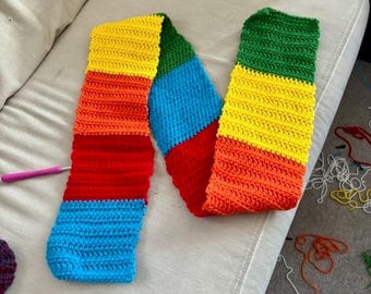 以下が含まれることがあります： 手作りの、虹色の模様のカラフルなかぎ針編みのスカーフ。赤、オレンジ、黄色、緑、青のセクションがあります。ピンクのかぎ針編みのフックが近くにあります。スカーフはニュートラルカラーの表面にあります。