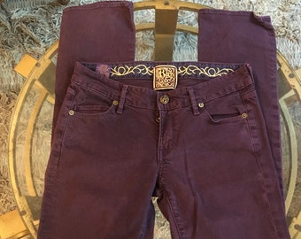 May include: Plum-colored denim jeans with a classic five-pocket design. The jeans feature a button and zipper closure, with a decorative label at the waistband. The jeans are laid flat on a glass table.