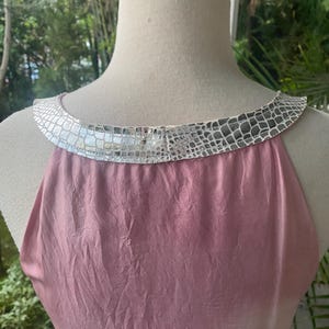 May include: A sleeveless top in a dusty rose pink color with a textured, crinkled fabric finish. The garment features a wide, rounded neckline trimmed with a metallic silver band that has a repeating, embossed reptile-like scale pattern.