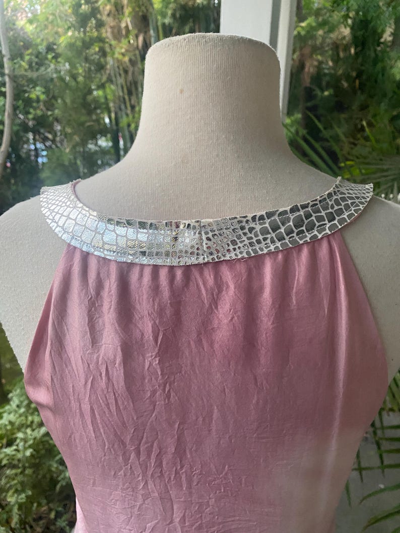 May include: A sleeveless top in a dusty rose pink color with a textured, crinkled fabric finish. The garment features a wide, rounded neckline trimmed with a metallic silver band that has a repeating, embossed reptile-like scale pattern.