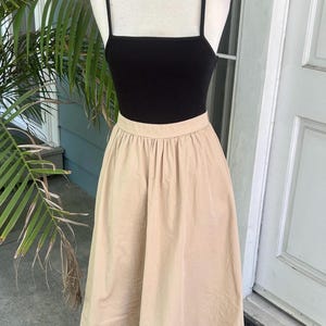 May include: A black ribbed tank top with thin straps paired with a beige midi skirt. The skirt has a gathered waist and falls to mid-calf. The outfit is displayed on a mannequin, suggesting a vintage or retro style.