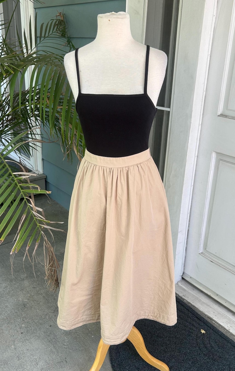 May include: A black ribbed tank top with thin straps paired with a beige midi skirt. The skirt has a gathered waist and falls to mid-calf. The outfit is displayed on a mannequin, suggesting a vintage or retro style.