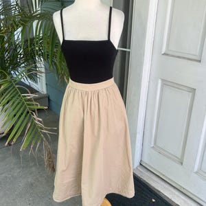 May include: A black ribbed tank top with thin straps is paired with a beige midi skirt. The skirt has a high waist and a flared silhouette. The outfit is displayed on a mannequin with a wooden stand, set against a backdrop of a house exterior.