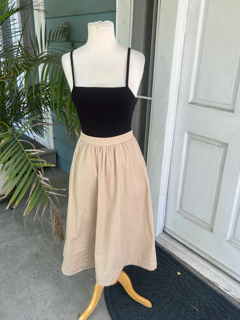 May include: A black ribbed tank top with thin straps is paired with a beige midi skirt. The skirt has a high waist and a flared silhouette. The outfit is displayed on a mannequin with a wooden stand, set against a backdrop of a house exterior.