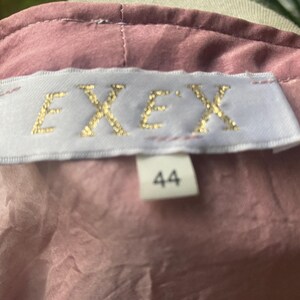 May include: A dusty rose-colored garment featuring a white rectangular fabric label sewn into the neckline. The label displays the text "EXEX" in gold lettering, with a smaller white tag below it showing the number "44" in black print.