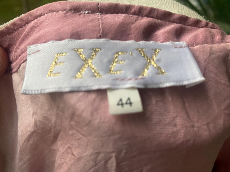 May include: A dusty rose-colored garment featuring a white rectangular fabric label sewn into the neckline. The label displays the text "EXEX" in gold lettering, with a smaller white tag below it showing the number "44" in black print.