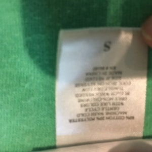 May include: A white clothing tag with the letter "S" at the top. The tag includes care instructions such as "Machine wash cold" and "gentle cycle". The tag is made of 80% cotton and 20% polyester.