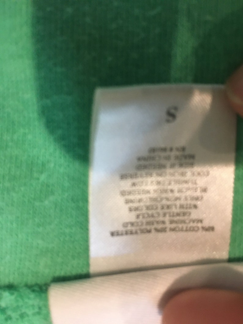 May include: A white clothing tag with the letter "S" at the top. The tag includes care instructions such as "Machine wash cold" and "gentle cycle". The tag is made of 80% cotton and 20% polyester.